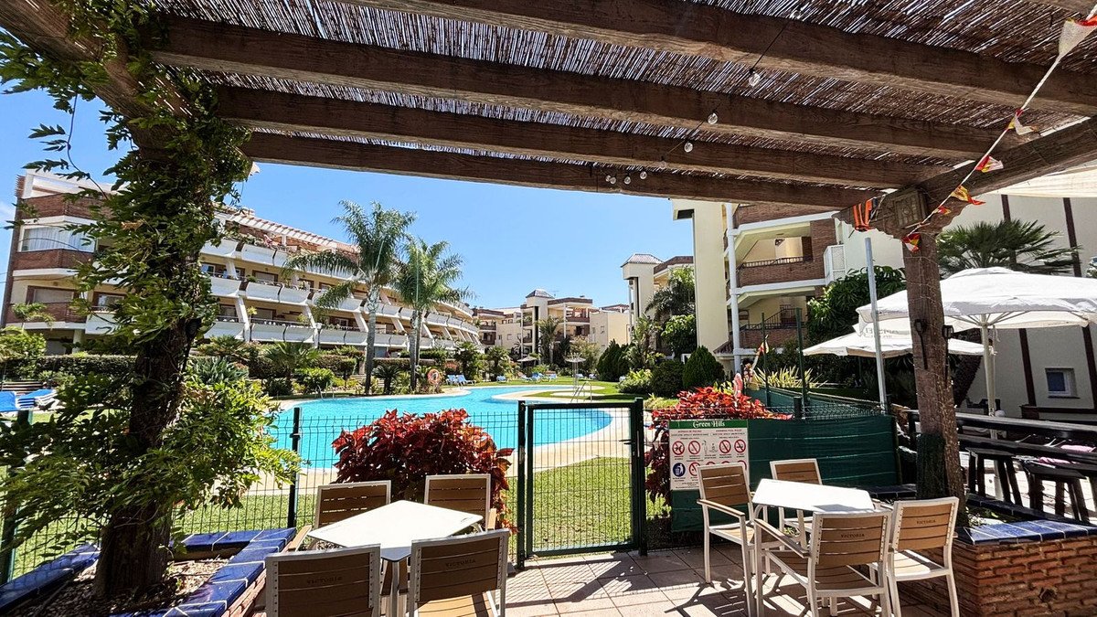 Outdoor terrace with tables and chairs overlooking a swimming pool and apartment buildings under a clear blue sky.
