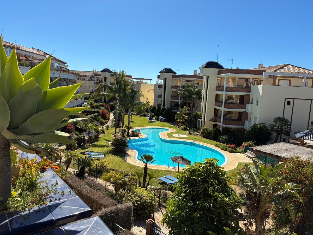 Resort complex with a large blue swimming pool, lush gardens, and modern apartment buildings under a clear blue sky.