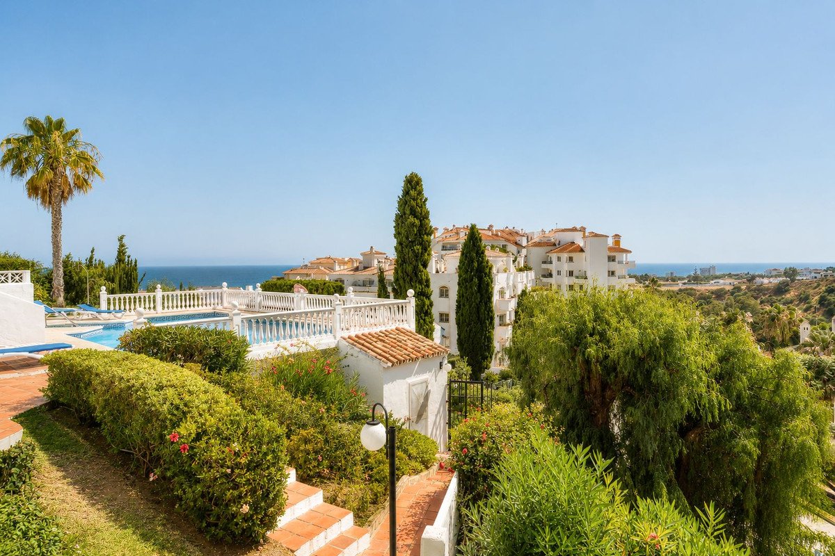 View of a swimming pool and terrace with lush greenery and distant sea views, residential buildings in the background.