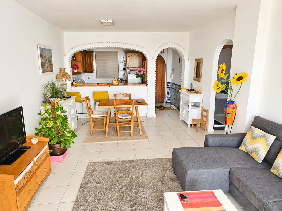 Spacious living and dining area with an open kitchen, featuring a sofa, dining table, and access to a balcony with sunflowers.