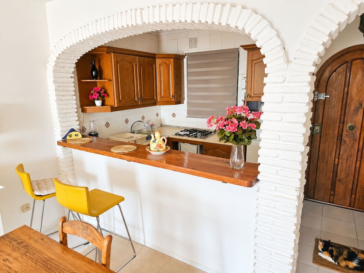 A cozy kitchen with wooden cabinets, a breakfast bar, yellow stools, and flowers. A cat rests by the door.