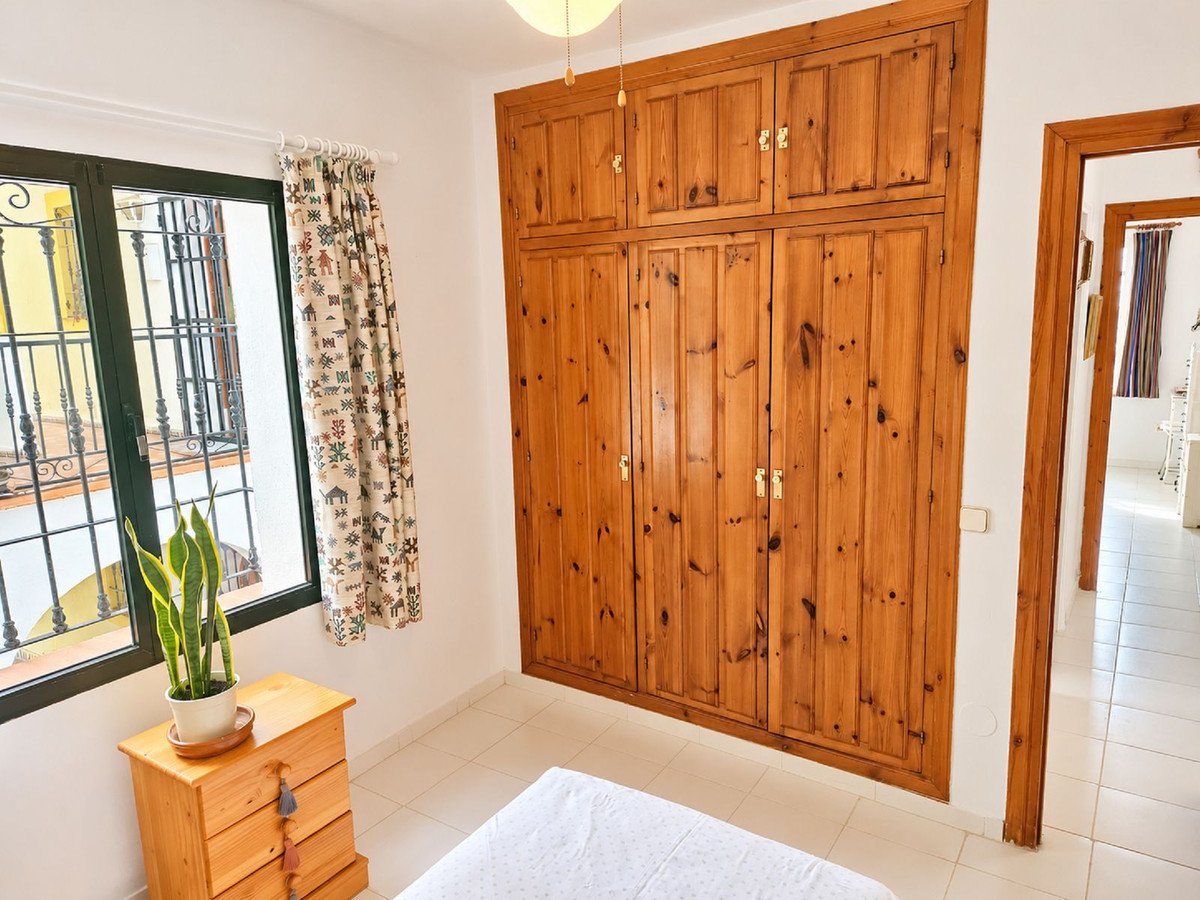 A bedroom with a large wooden wardrobe, a window with patterned curtains, and a small bedside table with a plant.