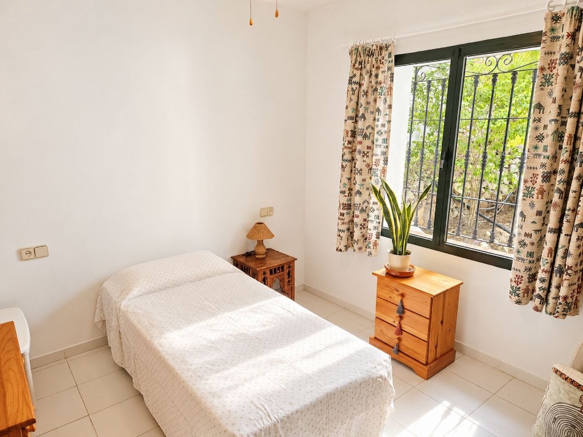 Bedroom with a single bed, a bedside table, a lamp, and a window with patterned curtains showing greenery outside.
