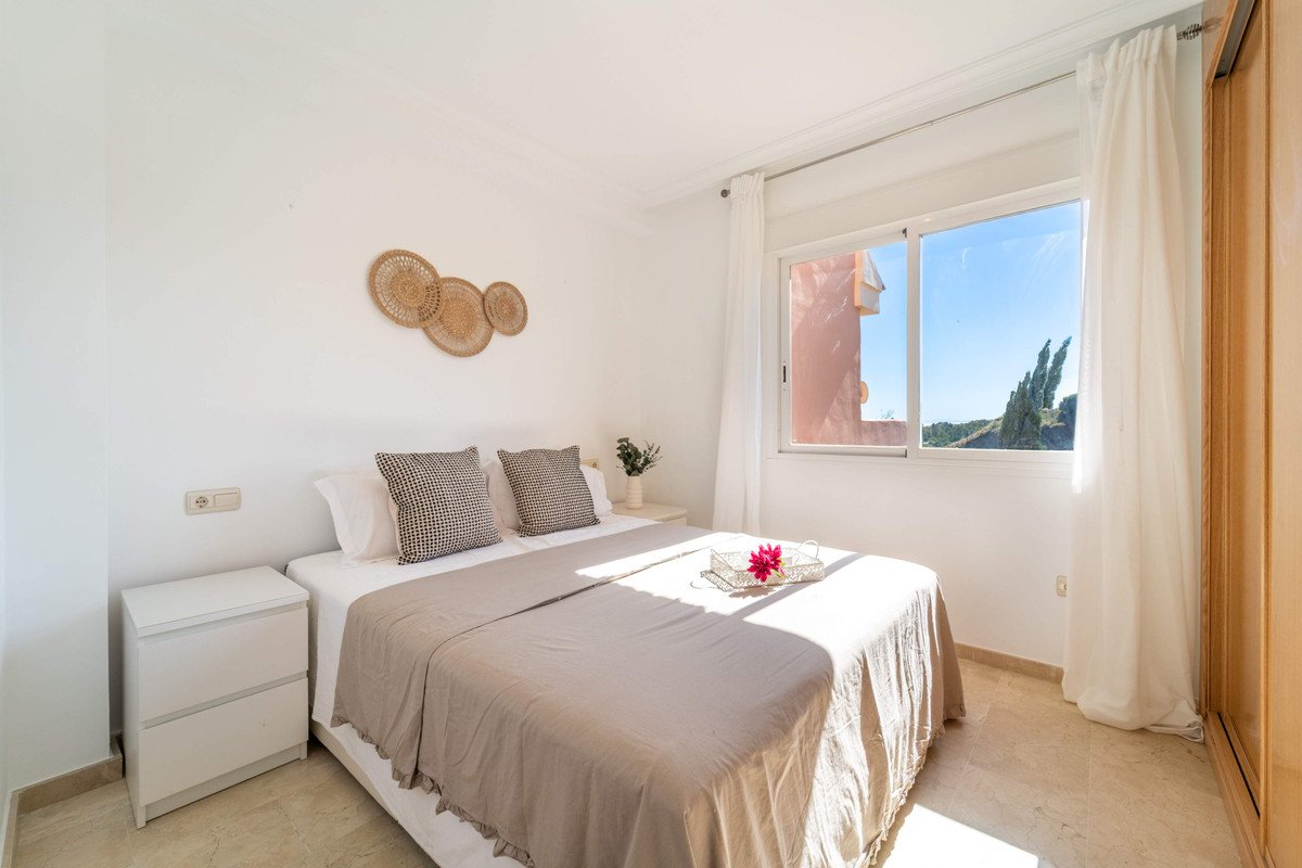 A bright bedroom with a king-size bed, bedside tables, and a large window offering a view of trees and buildings under a clear blue sky.