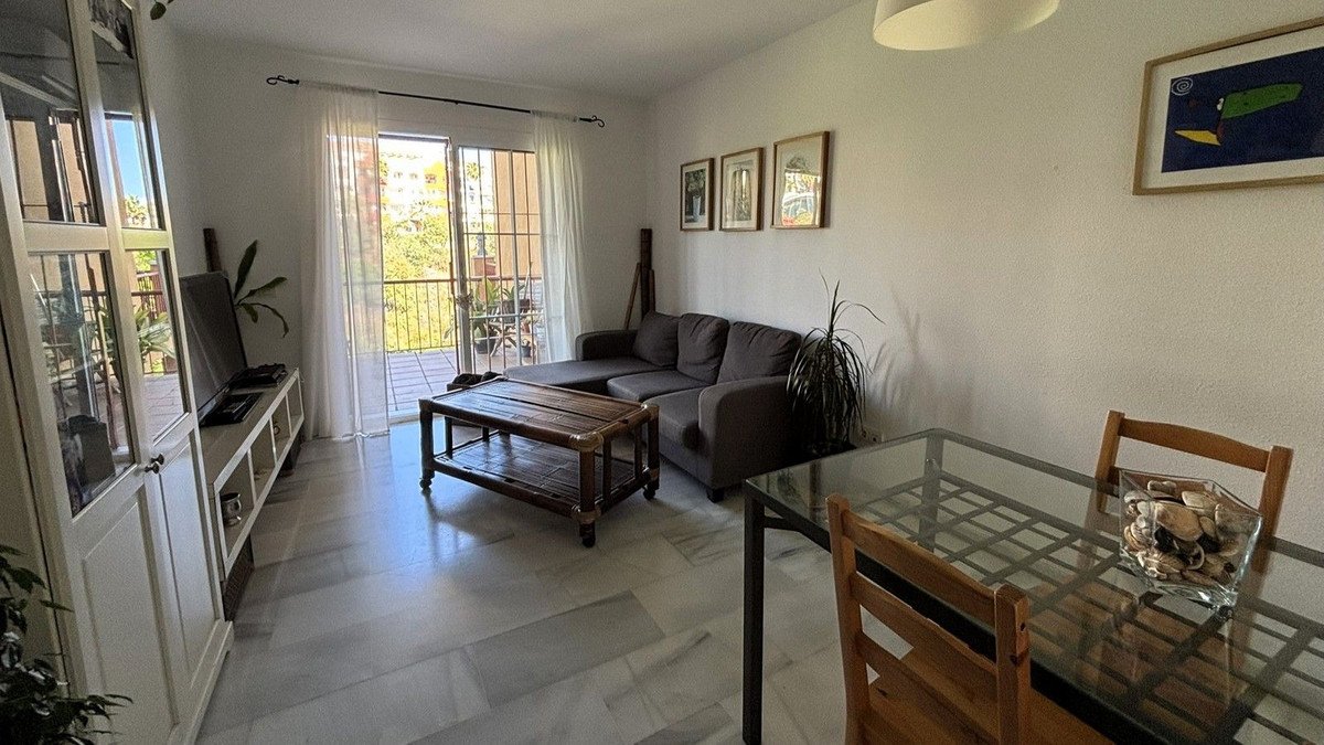A living and dining area with a sofa, coffee table, dining set, and a view of the balcony with greenery.