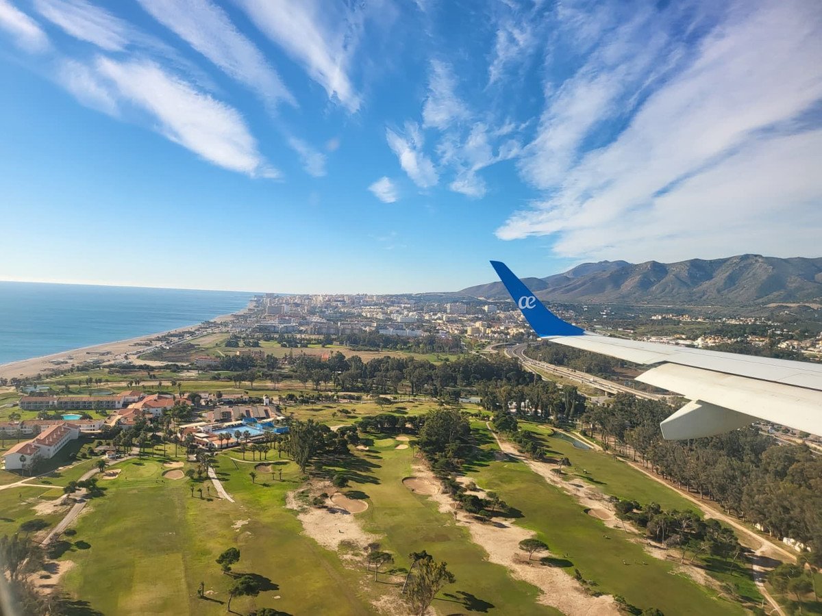 Aerial view of a coastal town with a golf course, beach, and mountains under a blue sky with clouds.