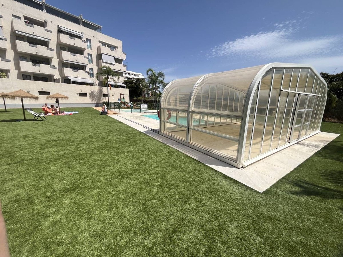 A modern apartment building with a covered swimming pool and a manicured lawn on a sunny day.