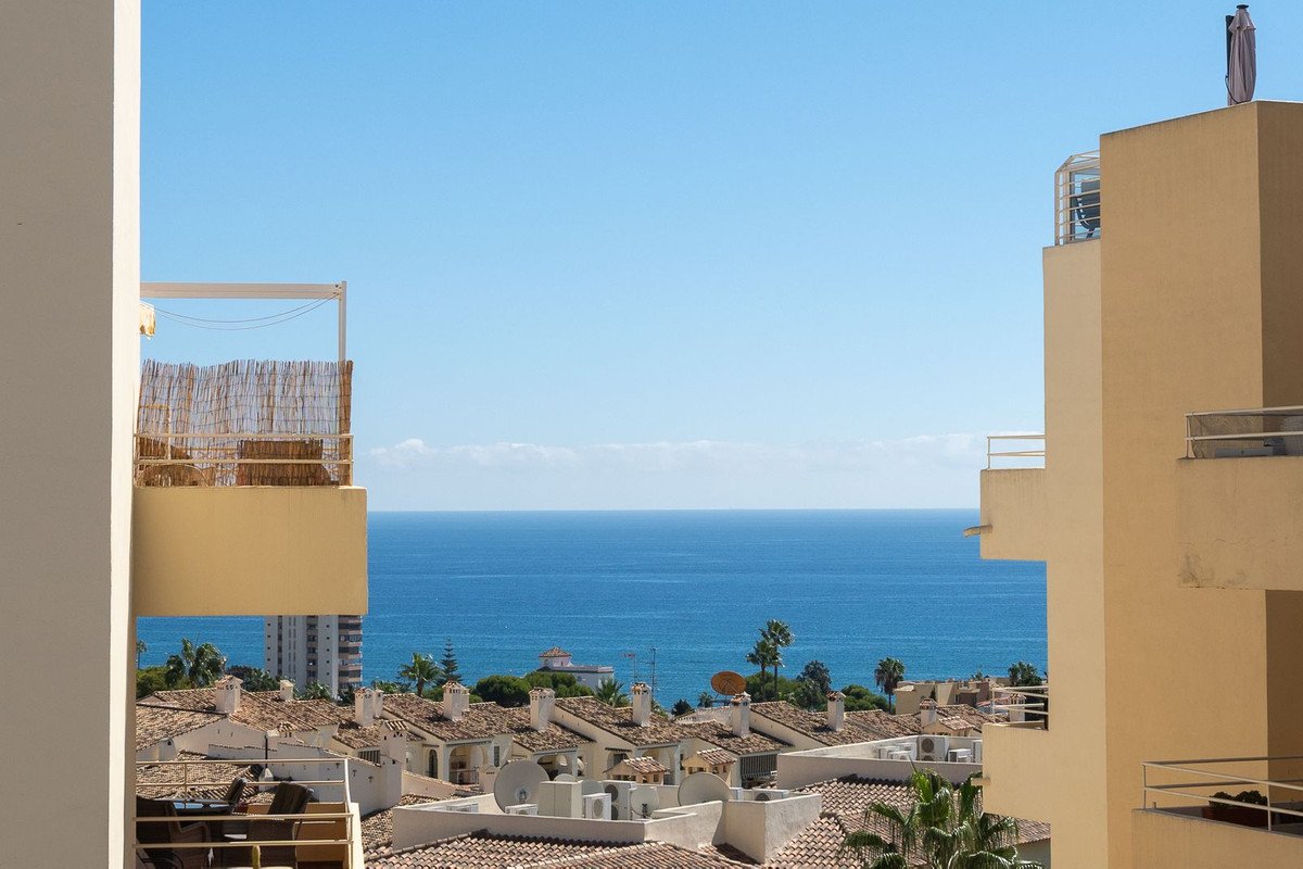 Expansive view of the blue sea from a balcony, overlooking rooftops and palm trees under a clear sky.