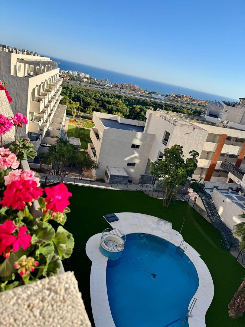 View from a balcony overlooking a swimming pool, artificial grass area, and the distant sea under a clear blue sky.