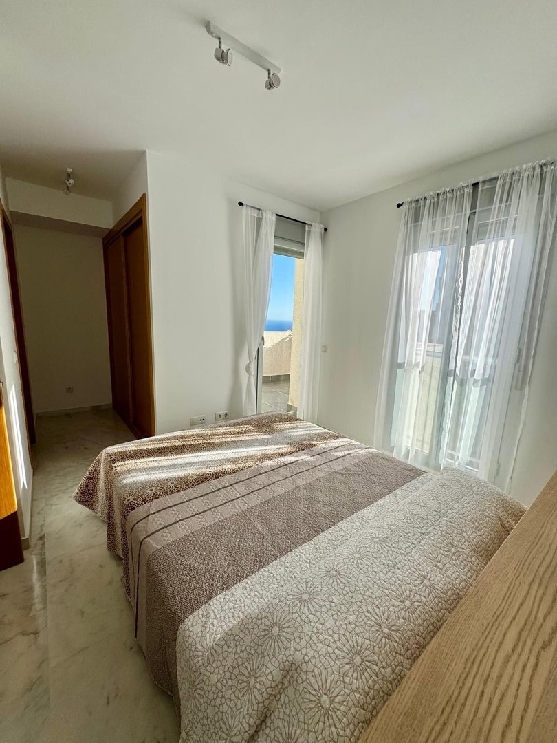 A bedroom with a double bed, patterned duvet, white sheer curtains, and a balcony with a sea view.