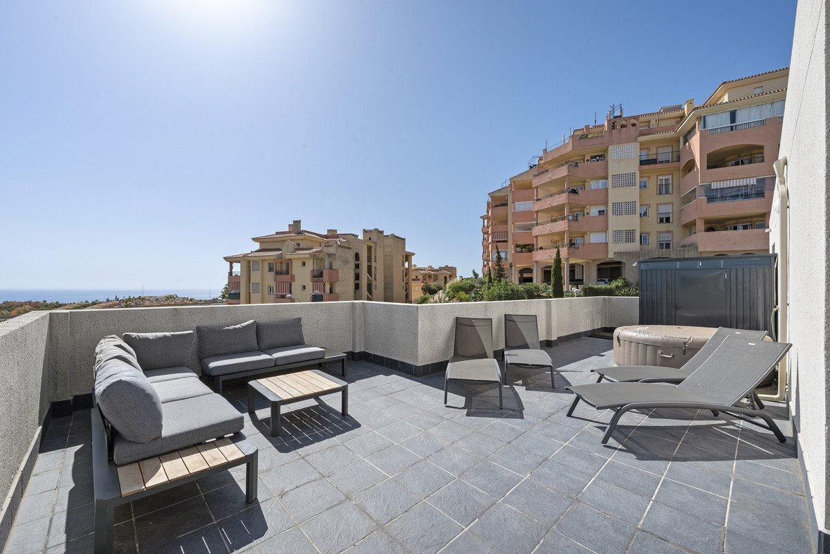Spacious terrace with outdoor sofa, coffee table, lounge chairs, and hot tub overlooking the sea and buildings.