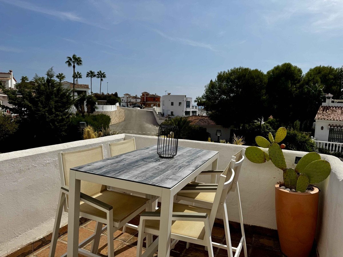 Outdoor dining area with a table and chairs on a terrace, overlooking a street with palm trees and houses.