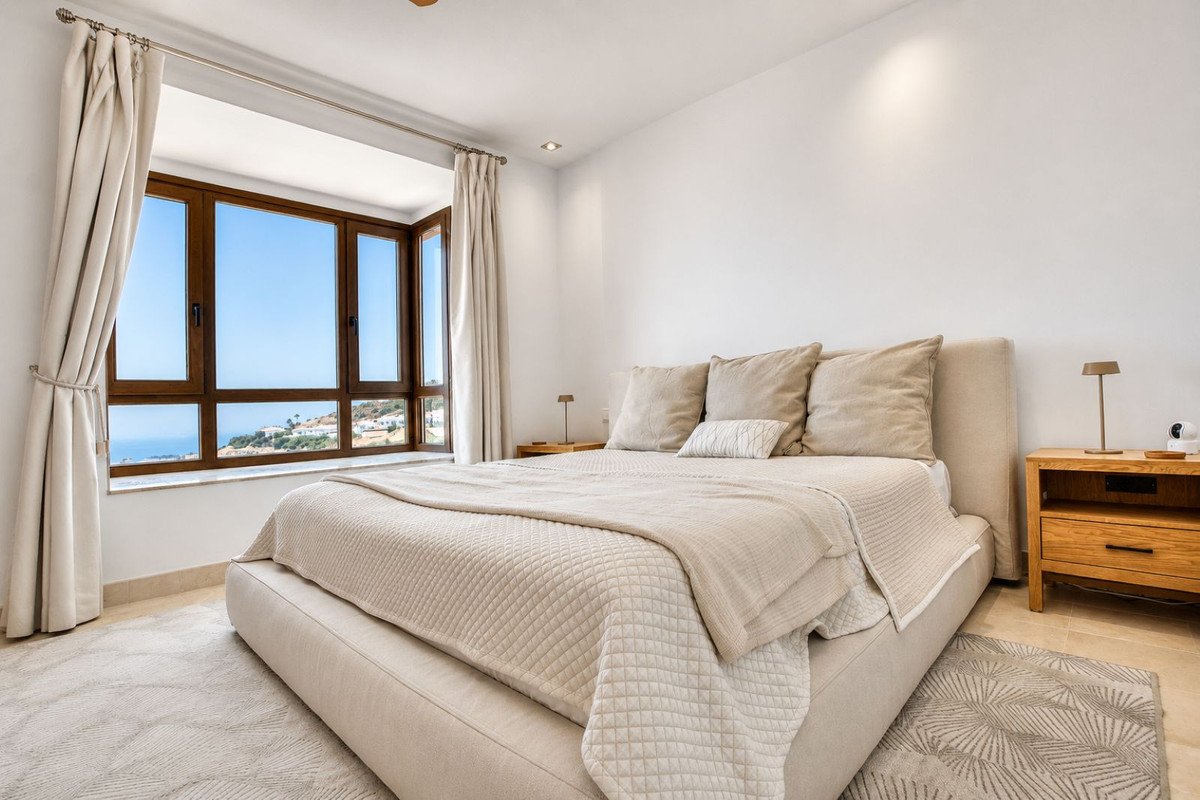 Modern bedroom with a large bed and a bay window offering panoramic views of the sea and coastal town.