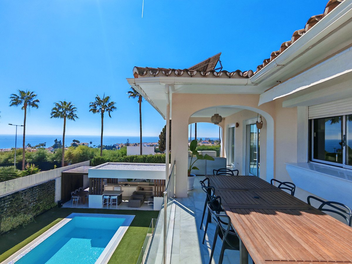 Spacious terrace with dining table overlooking a swimming pool, outdoor kitchen, and the sea with palm trees.