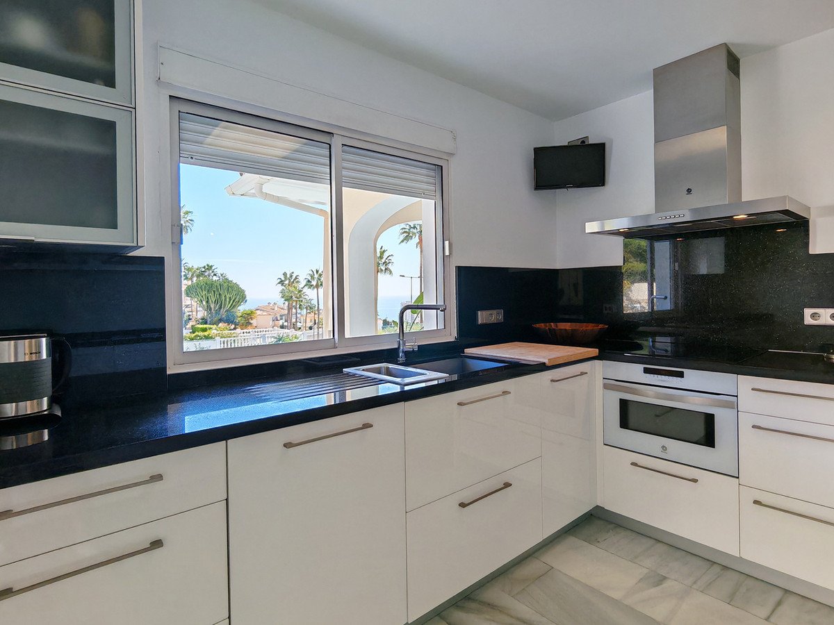 Modern kitchen with white cabinets, black countertops, and a window overlooking palm trees and the sea.