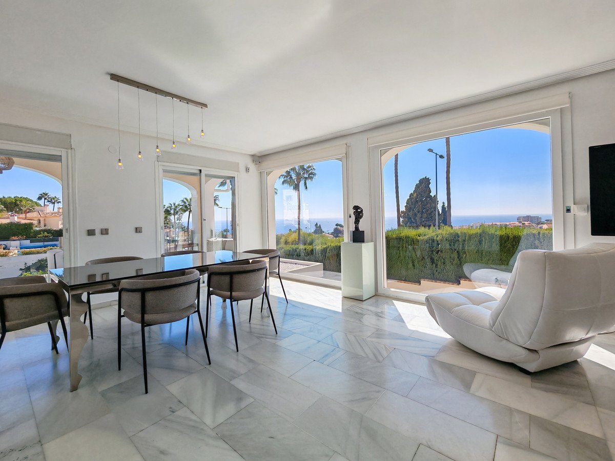 Modern dining room with large windows overlooking the sea, palm trees, and a plush white armchair.