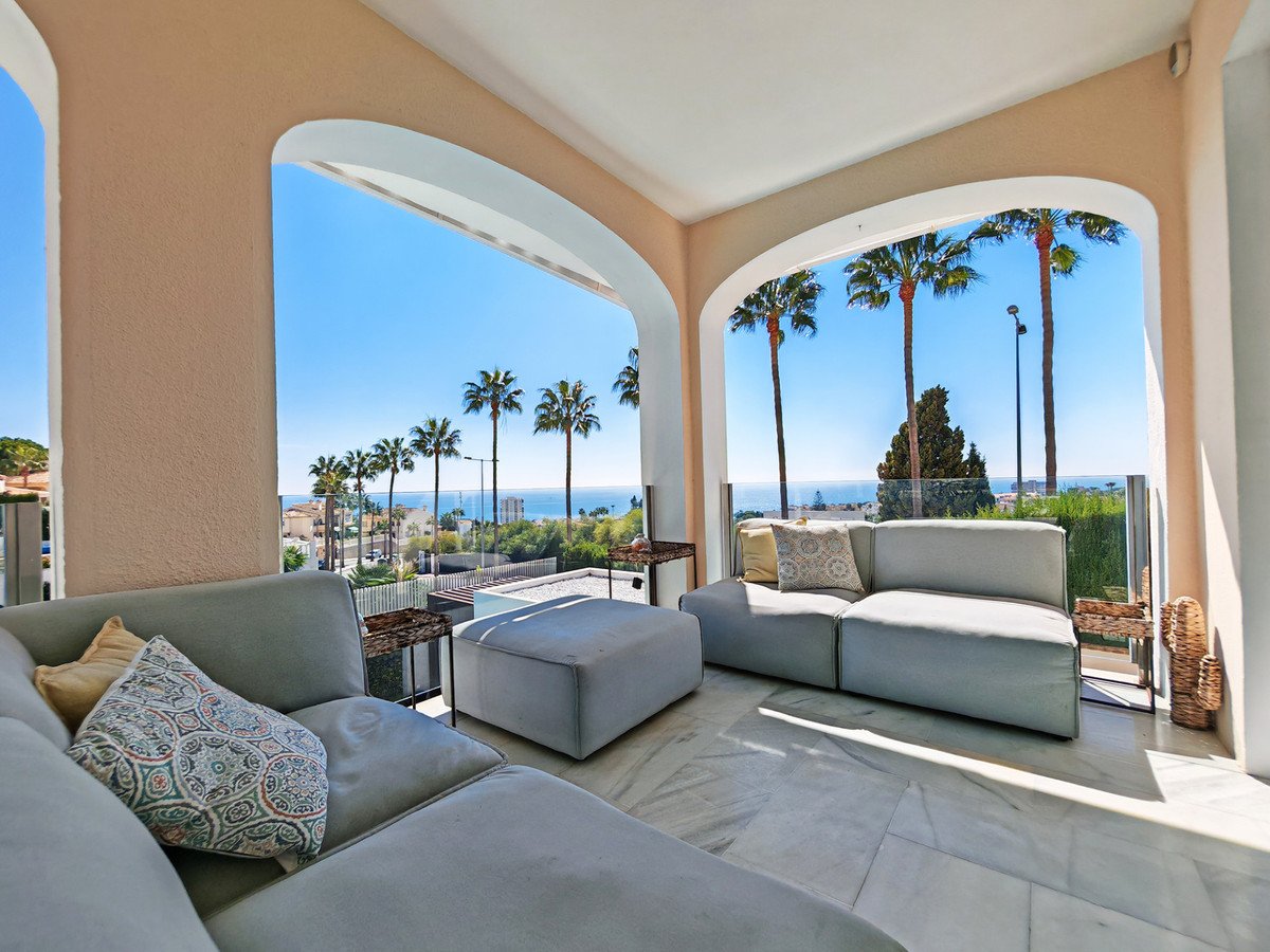 Covered terrace with comfortable seating, offering stunning sea views framed by palm trees.