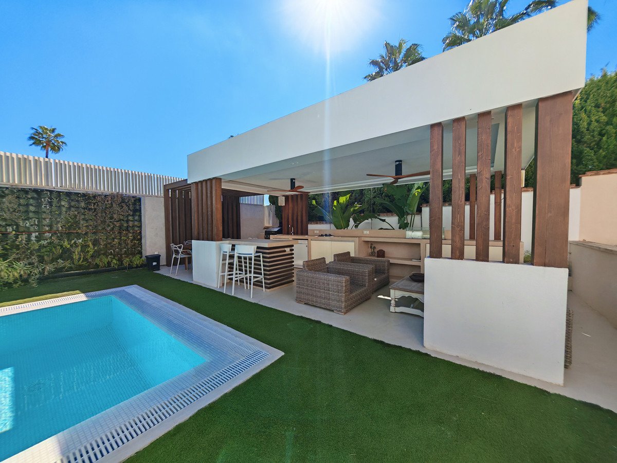 Modern outdoor kitchen with bar seating, lounge area, and a swimming pool on a sunny day.