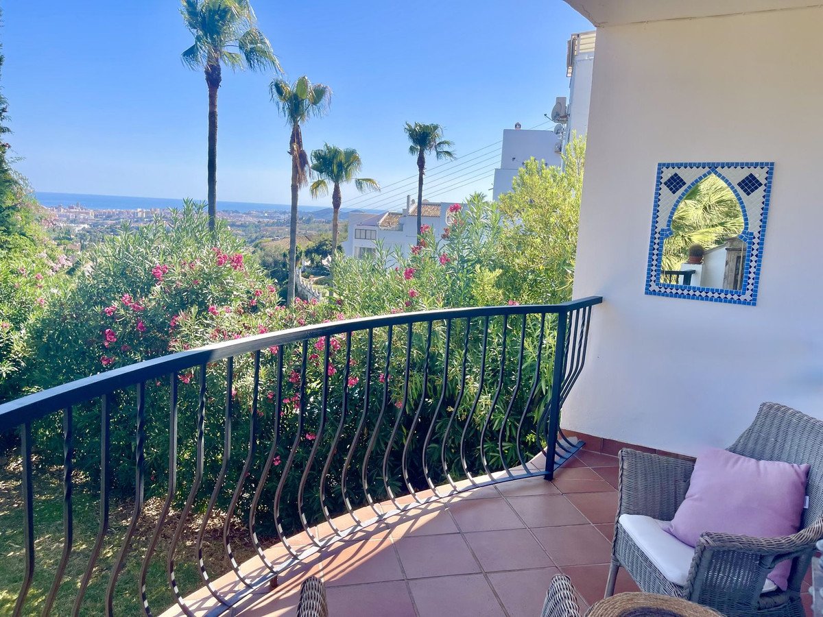 Terrace with ornate mirror and seating, overlooking lush greenery and a distant coastal town with palm trees.