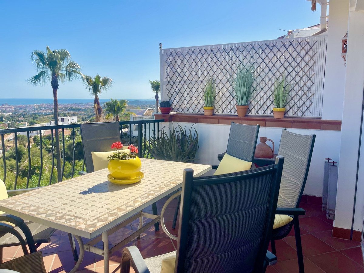 Terrace with dining table and chairs overlooking the sea and distant town, with palm trees.