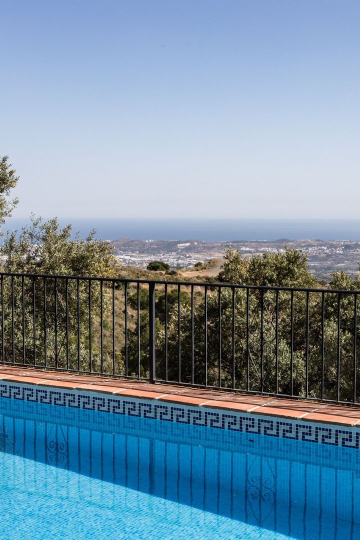 Blue swimming pool with a tiled border, overlooking a coastal town and the sea under a clear sky.