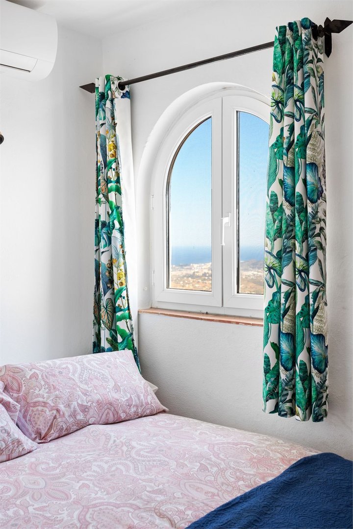 Bedroom with an arched window, decorative curtains, and a view of the sea and town.
