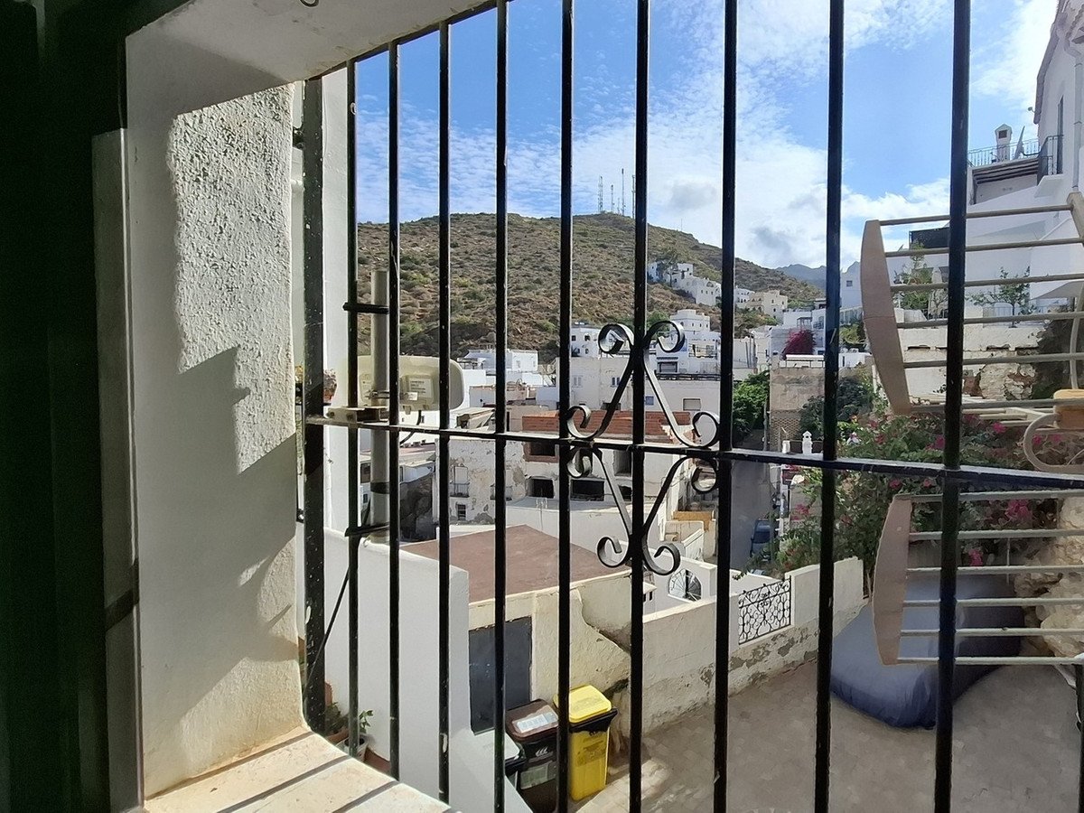 View from a window with bars, overlooking a village nestled against hilly terrain under a blue sky.