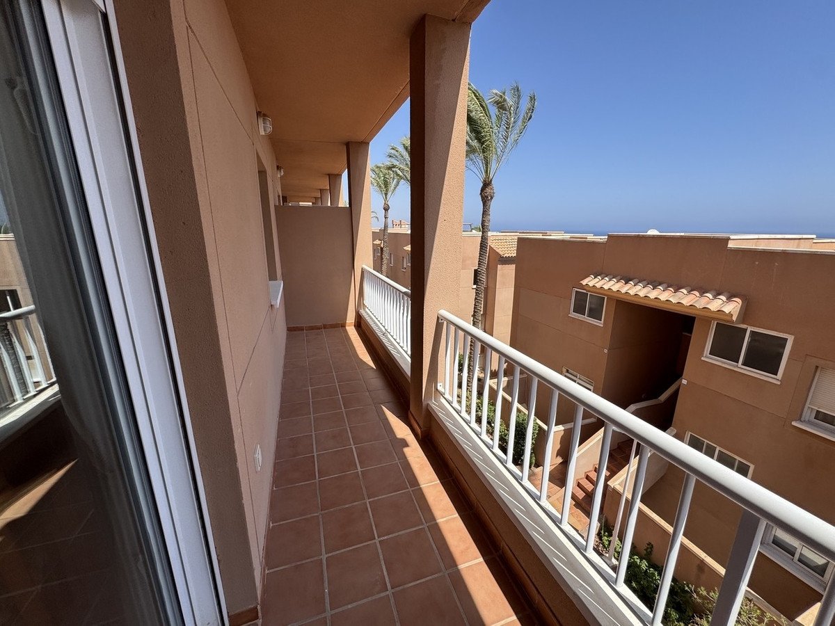 A long balcony with tiled floor and white railings overlooks a courtyard with palm trees and the distant sea under a clear blue sky.
