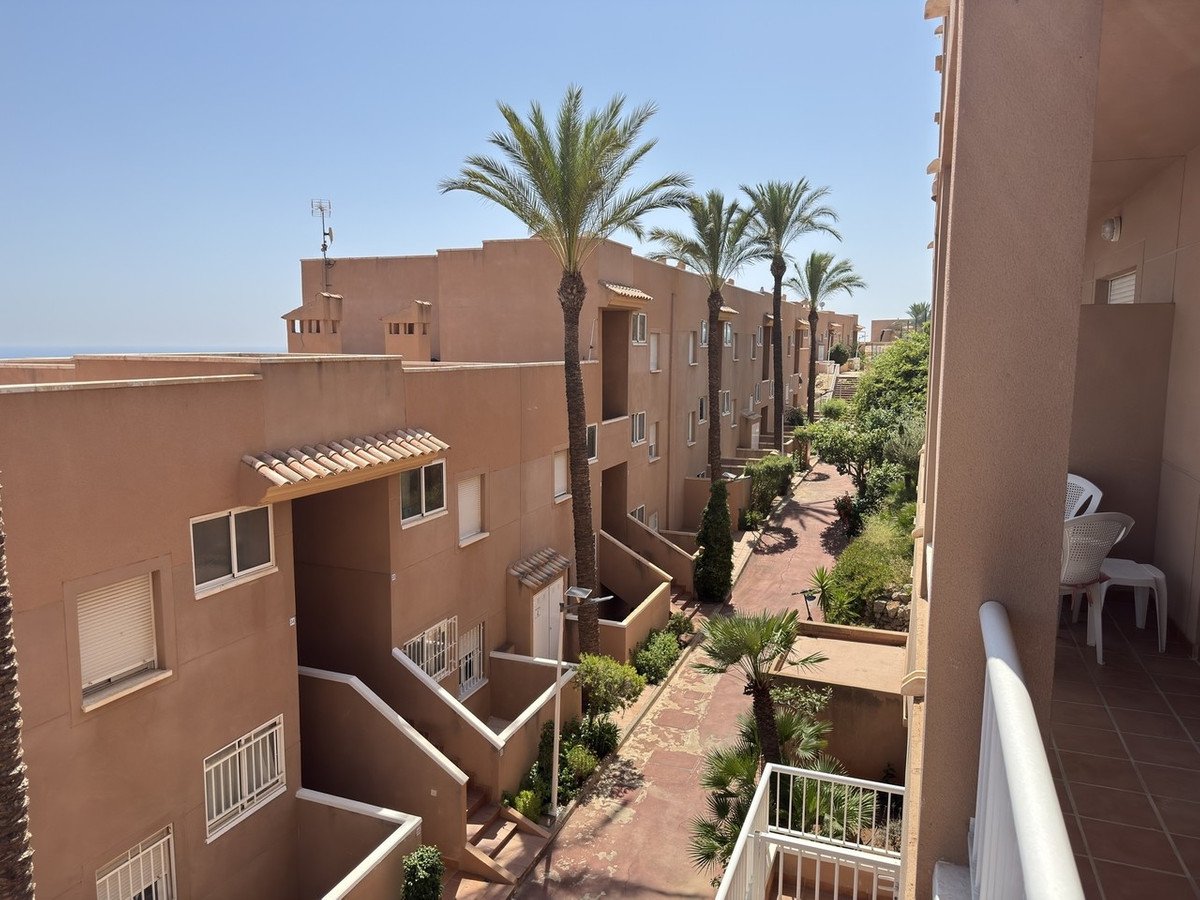 A view of a complex with apartment buildings and palm trees, overlooking the sea under a clear blue sky.
