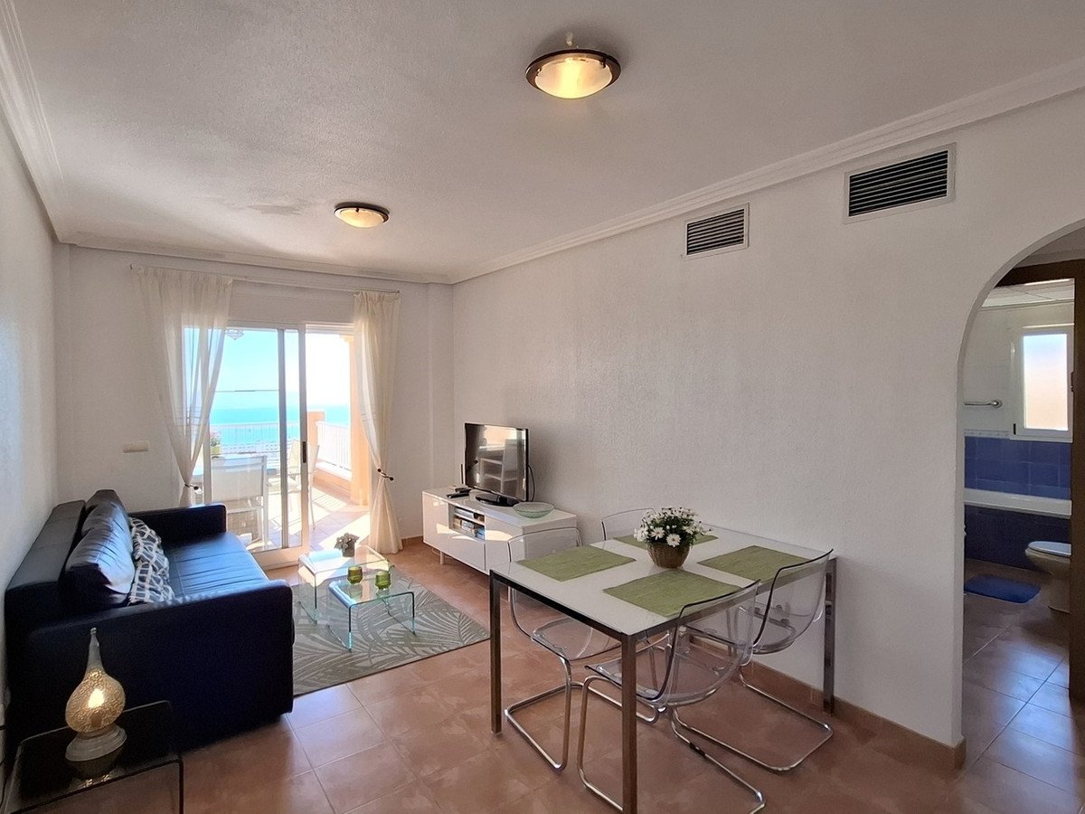 Modern living and dining area with a sofa, table, and balcony overlooking the sea. A bathroom is visible through an archway.