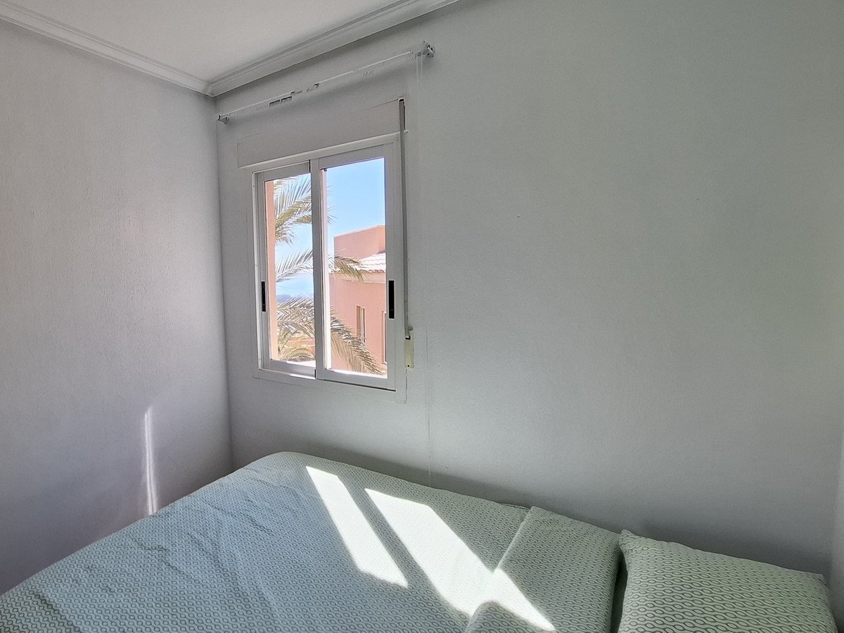 A simple bedroom with a bed and a window showing a view of palm trees, a building, and the sea under a blue sky.