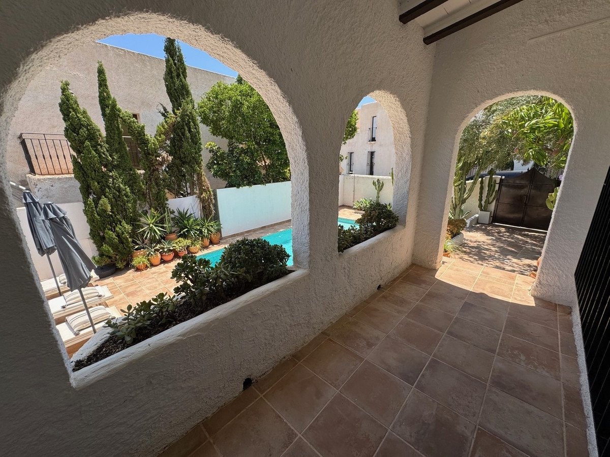 View through arched entryway to a patio with pool, lounge chairs, umbrellas, and lush landscaping on a sunny day.