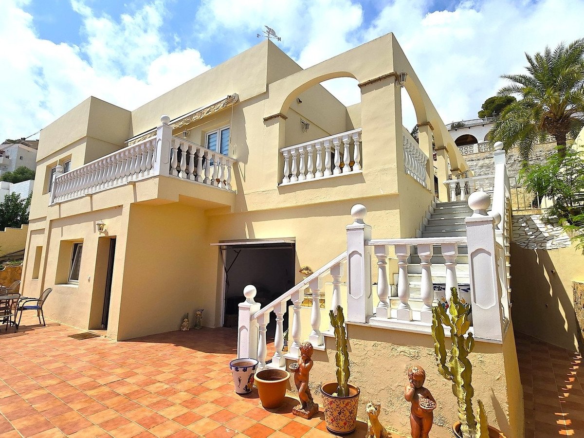 A multi-level house with white balustrades and arched features, set against a blue sky with clouds. Features a terrace and staircase.