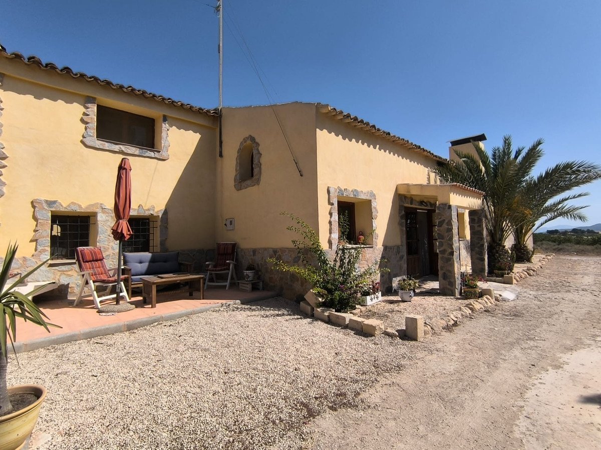 A yellow country house with a terrace featuring seating, an umbrella, and palm trees, set against a clear blue sky.