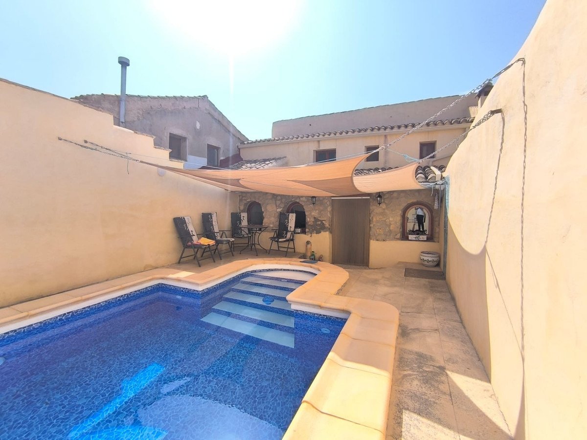 A private swimming pool with steps and a patio area with chairs under a shade sail on a sunny day.