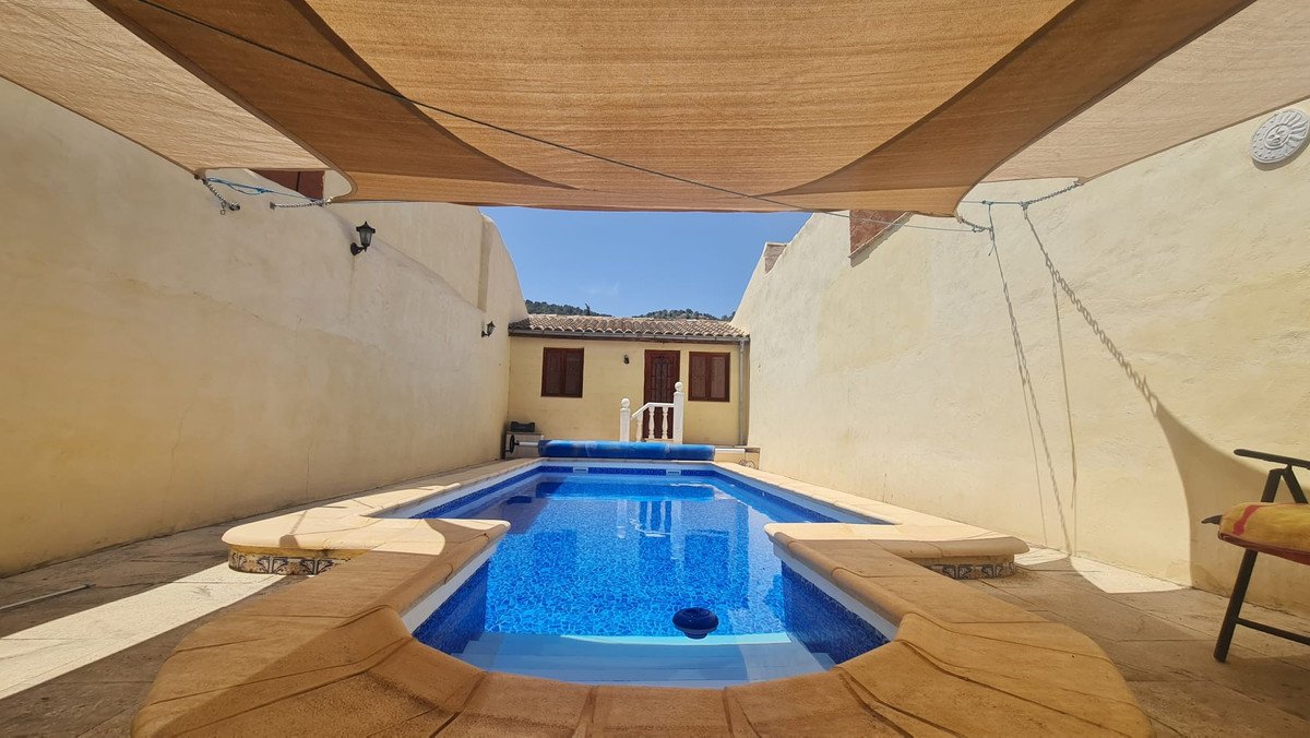 Rectangular swimming pool with blue water, surrounded by a tiled terrace and shaded by a canopy. A small house is visible in the background.