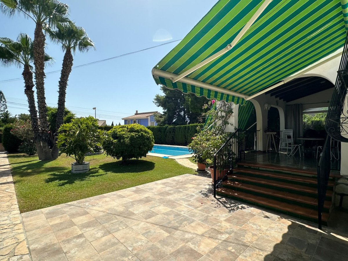 Backyard with a swimming pool, tiled patio, and green striped awning over a covered seating area with palm trees.