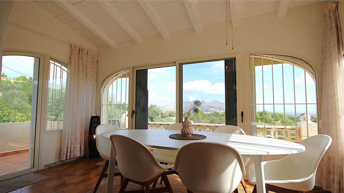 Modern dining room with white table and chairs, featuring large windows with a balcony and stunning mountain views.