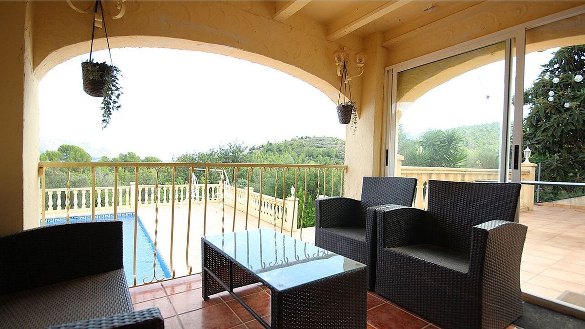 Covered patio with rattan furniture overlooking a swimming pool and lush green hills.