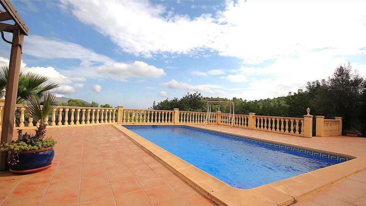 A rectangular swimming pool on a tiled patio with a balcony overlooking green hills and a cloudy sky.