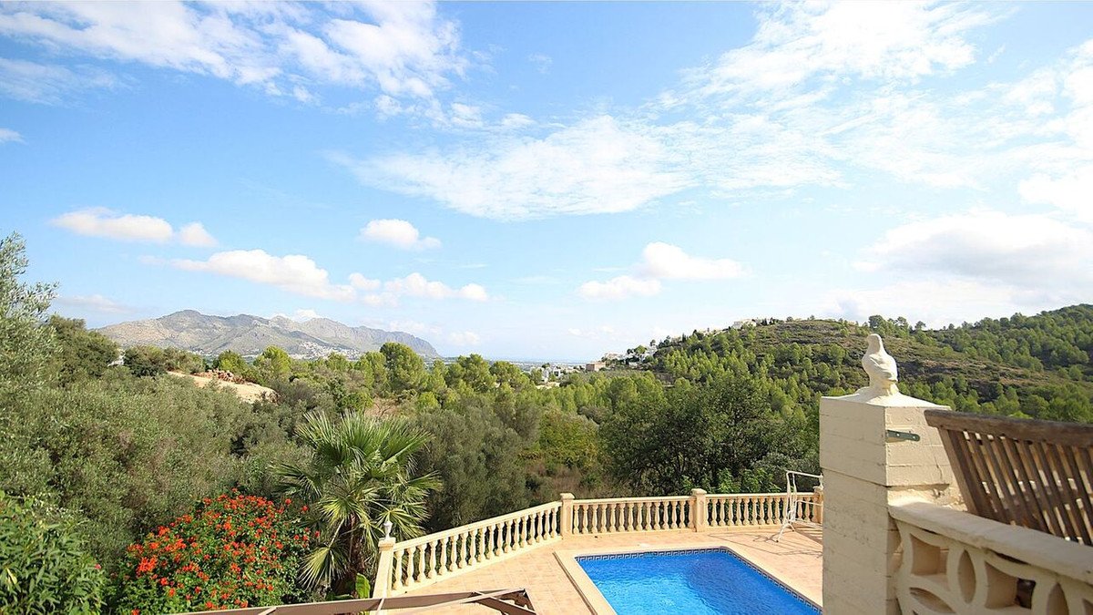 View of a swimming pool and a green landscape with mountains in the background under a cloudy blue sky.