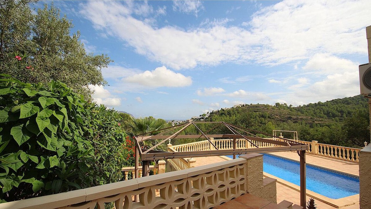 Terrace with a swimming pool and pergola, overlooking a green hill landscape under a blue sky with clouds.