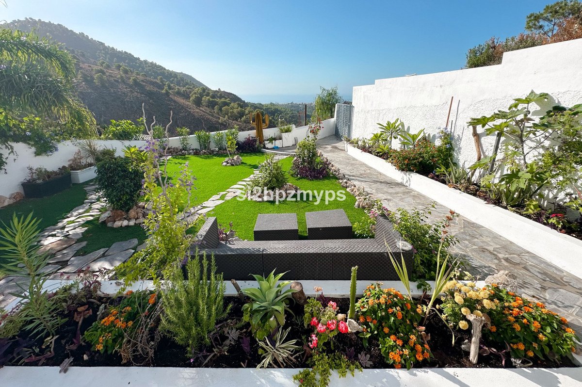 Manicured garden with artificial turf, stone pathways, flowering plants, and outdoor seating, overlooking a distant sea view.