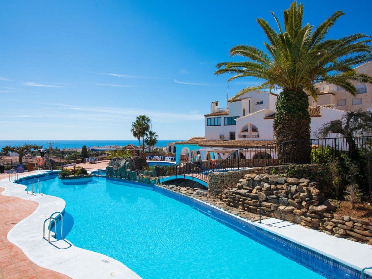 A large, bright blue swimming pool with a palm tree and a white resort building overlooking the sea on a sunny day.