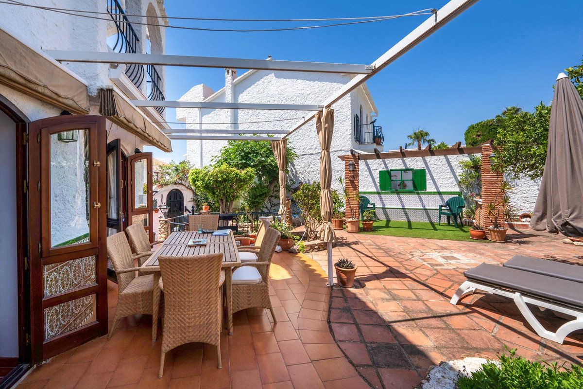Outdoor dining area with wicker chairs and wooden table on tiled patio, leading to a green garden with sun loungers and umbrella.
