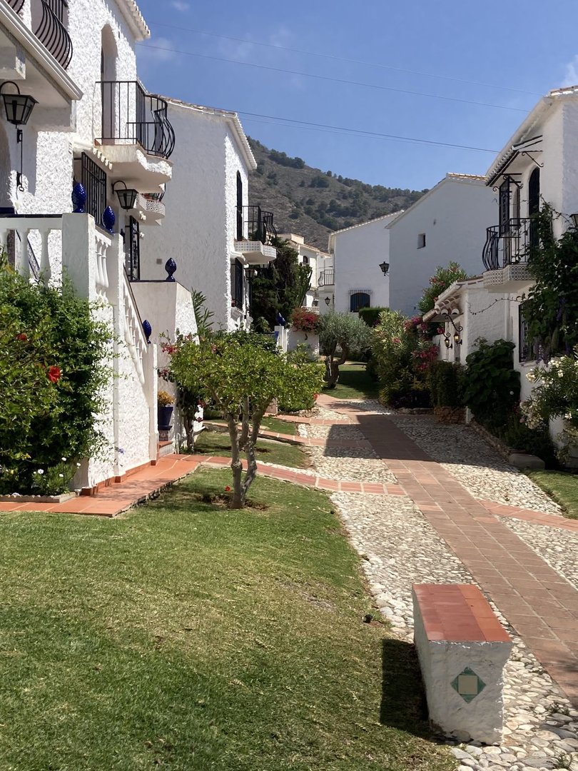 Charming white villas with balconies, pathways, green lawn, and lush vegetation leading towards hills.