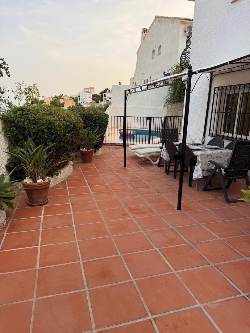 A tiled terrace with a dining set and lounge chairs overlooking a small pool and distant houses.