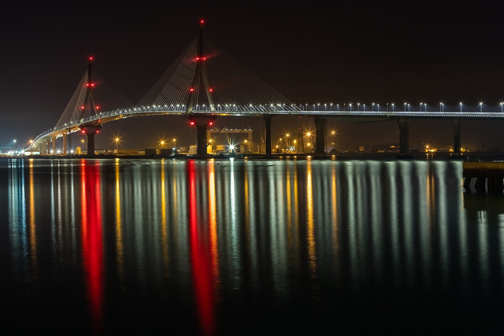 Alt text: Night view of a modern cable-stayed bridge with city lights reflecting on calm water.