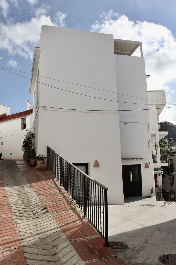 A white building with a terrace on top, seen from a sloped tiled path with a metal railing.