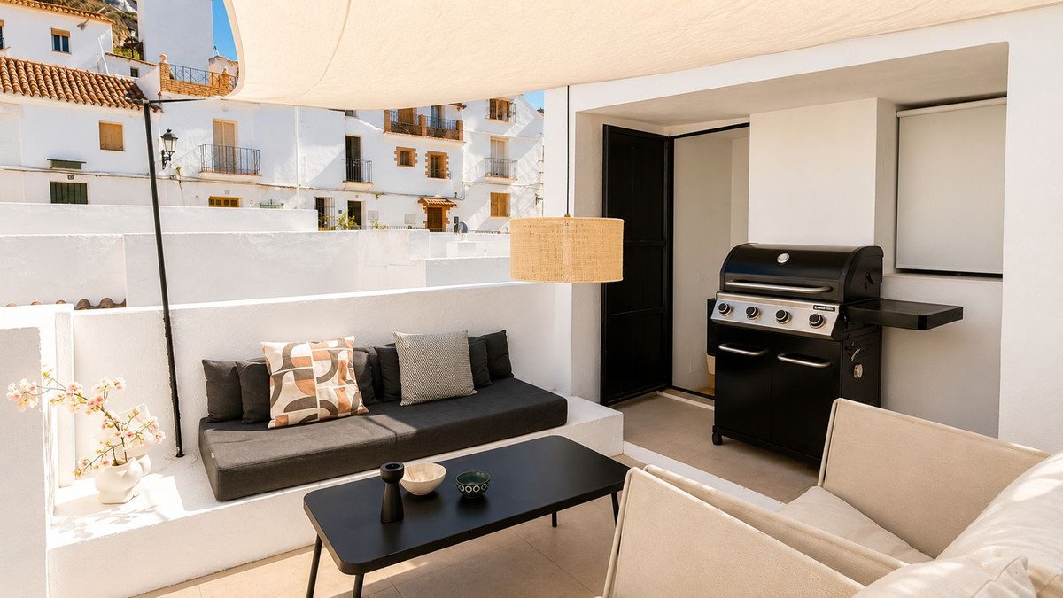 A rooftop terrace with built-in seating, a grill, a coffee table, and a view of white buildings.